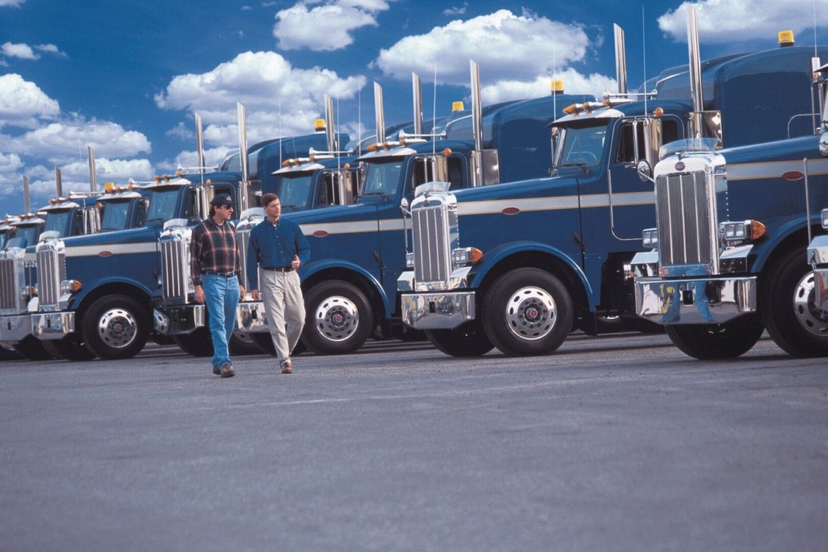 Two men walk and talk in front of a row of parked blue semi-trucks under a partly cloudy sky.