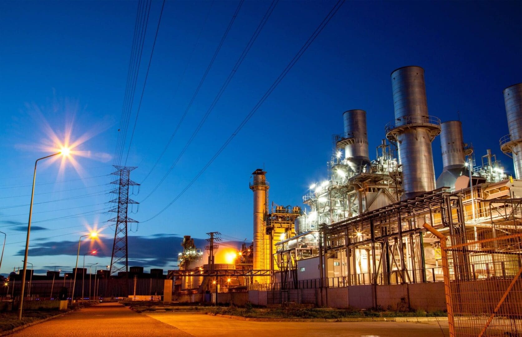 An industrial facility with tall chimneys and illuminated infrastructure at dusk, under a clear blue sky. Streetlights and power lines are visible in the foreground.