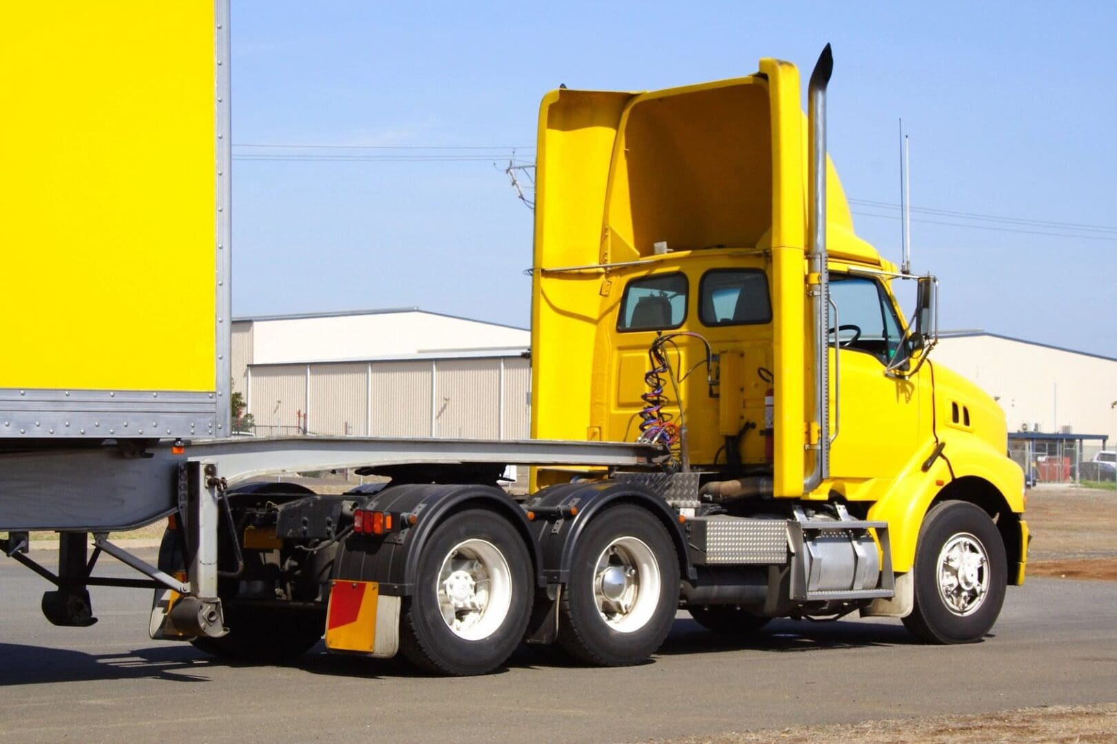 A yellow semi-truck with its cab hood opened, parked on a paved surface with industrial buildings visible in the background.