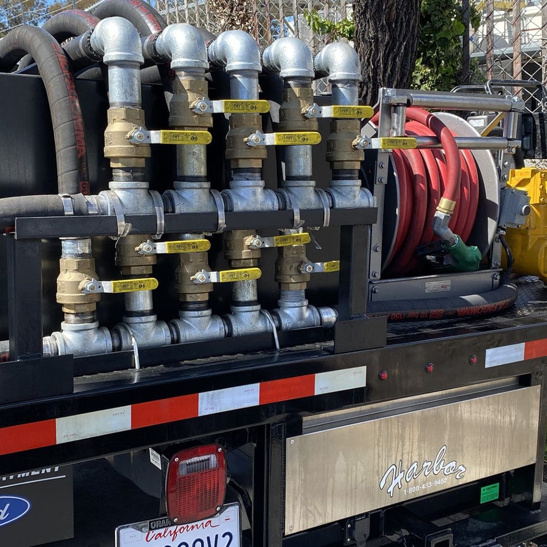 A close-up of industrial machinery featuring a row of metal pipes, valves, and red hoses mounted on a truck. A sign on the truck reads "Harbor." Partial view of a license plate at the bottom.