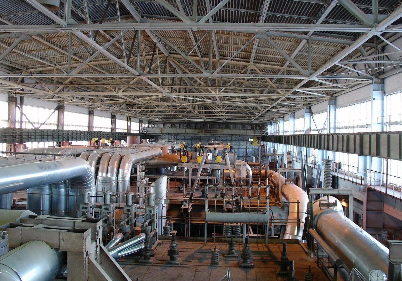 Interior of an industrial facility with a complex network of large metal pipes and beams, and workers in yellow safety gear visible on platforms in the background.